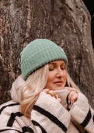 A person with closed eyes, wearing the warm and cozy Mist Chunky Beanie by Mimi & August and a striped sweater, stands against a tree, holding the collar close to their face.