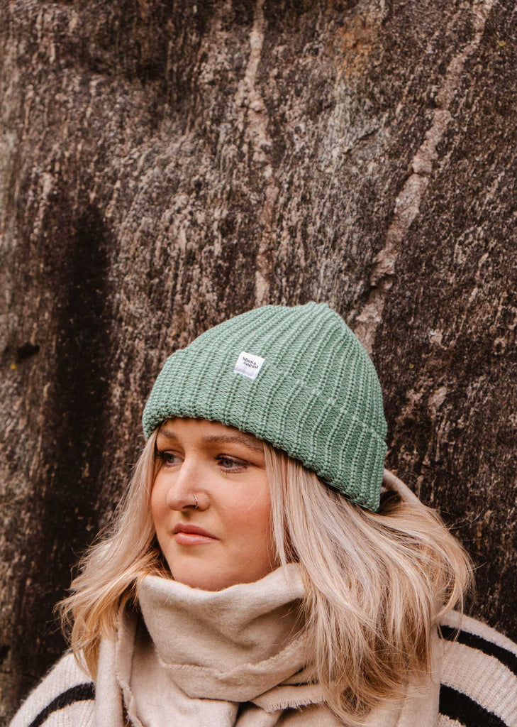 A person adorned in the winter essential Mist Chunky Beanie by Mimi & August, sporting a soft muted mist color along with a matching scarf, stands before a textured rock surface.