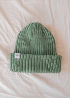 A green knitted beanie, the Mist Chunky Beanie by Mimi & August, is displayed on a white fabric background. This winter essential features a folded brim with a small white tag attached and comes in a soft muted mist color.