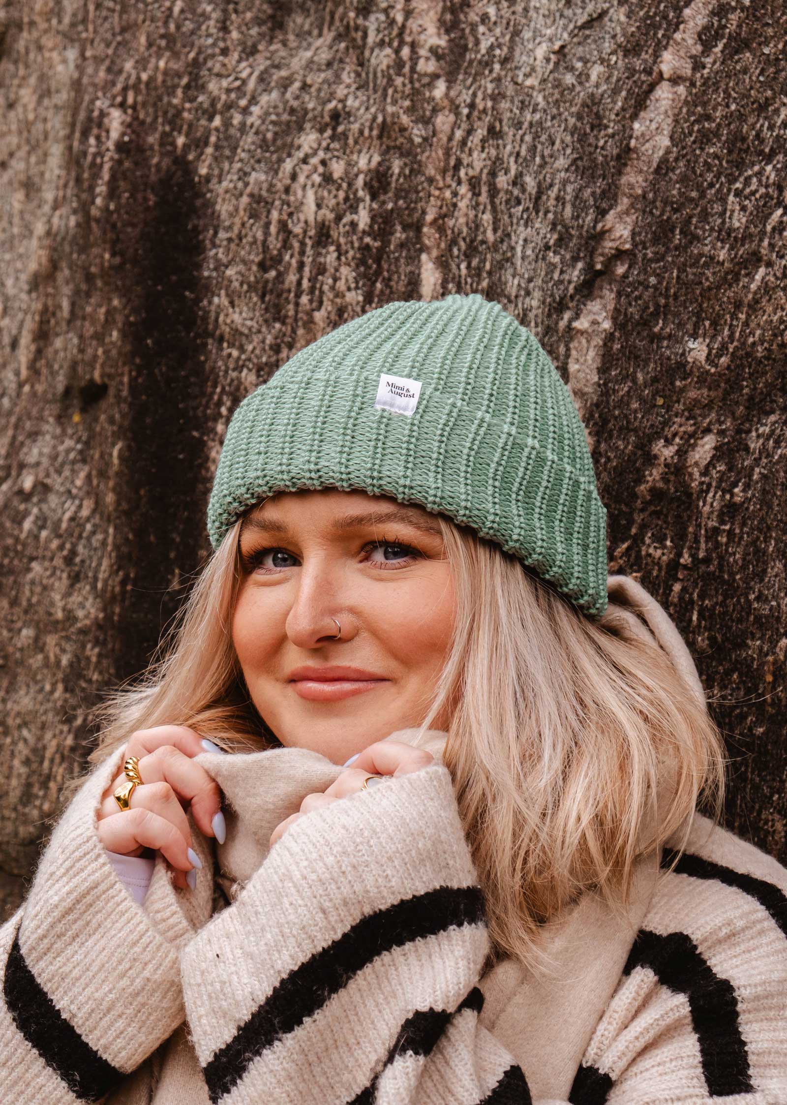 Une femme portant un bonnet vert Mist Chunky Beanie de Mimi & August et un pull rayé se tient à l'extérieur, souriant légèrement tout en tenant son col contre un grand rocher texturé en arrière-plan.