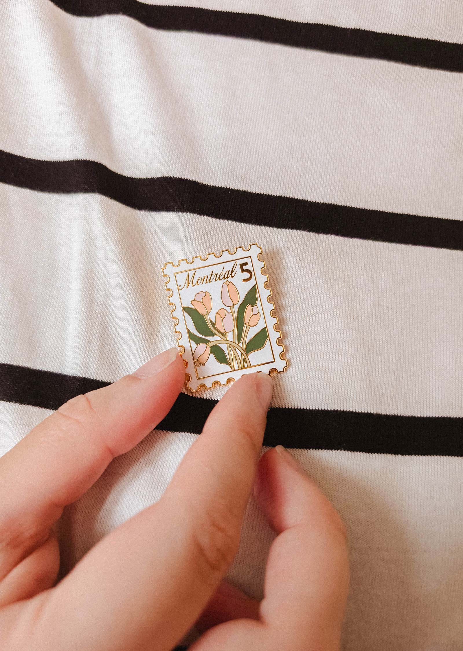 A hand holds the Montréal Stamp Enamel Pin by Mimi & August—an enamel pin styled like a postage stamp with tulips and "Montreal 5" text—set against a black and white striped fabric background. Perfect for flower lovers.
