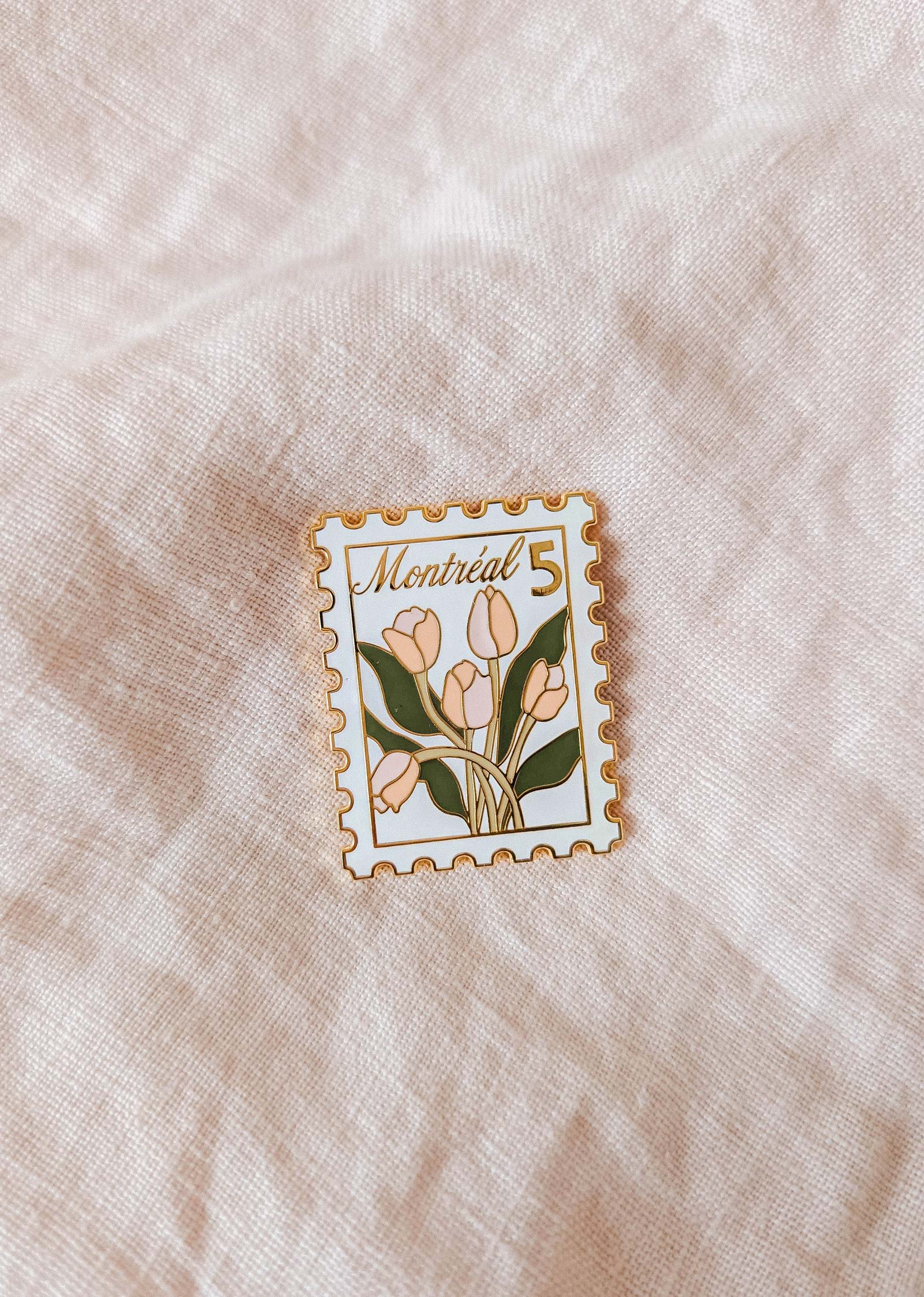 The Montréal Stamp Enamel Pin by Mimi & August is a tulip-themed, stamp-shaped enamel pin with “Montréal 5” text—a charming accessory for flower lovers. Shown on light fabric.