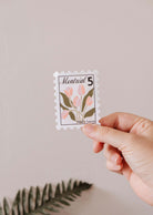Une main tient le timbre Montréal - Autocollant en vinyle de Mimi & August, avec des fleurs roses, des feuilles vertes et "Montréal 5" en haut sur un fond uni.
