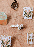 Five enamel pins from Mimi & August, including floral stamps, a squirrel, “Montréal Yo” enamel pin, “BAGEL MTL,” and “JARDIN DES ROSES,” are arranged on a wooden surface—each uniquely designed in Montreal.