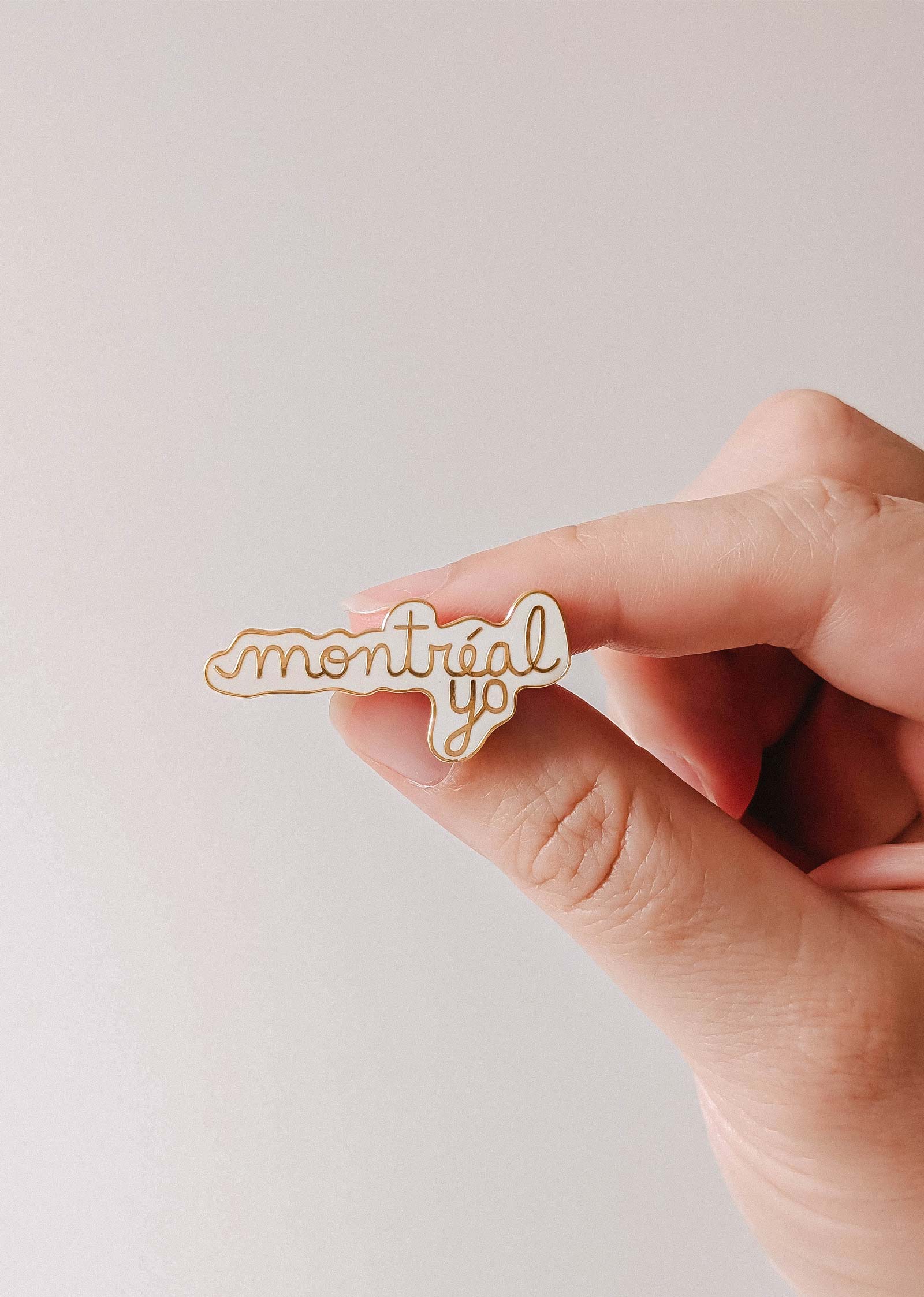 A hand holds the Montréal Yo Enamel Pin by Mimi & August, featuring "montréal yo" in cursive on a white background and crafted from gold tone brass enamel.