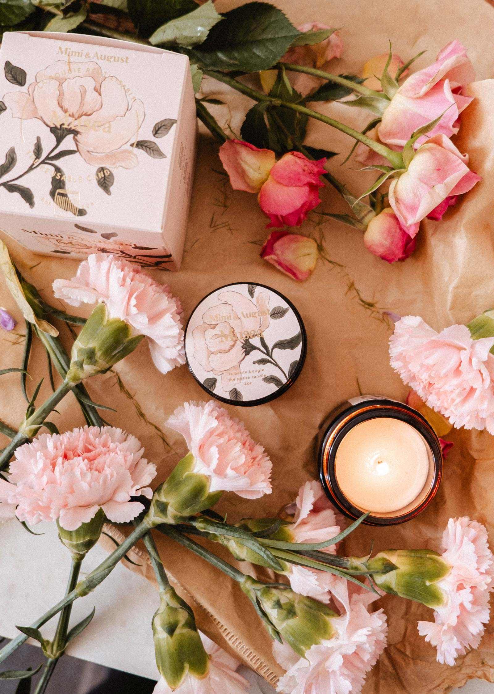 La bougie de soya Musea 2 oz de Mimi & August, une crème corporelle au parfum floral et une boîte décorative sont disposées sur du papier brun avec des œillets et des roses roses. Le contenant en verre réutilisable ajoute une touche écoresponsable à cet ensemble élégant.