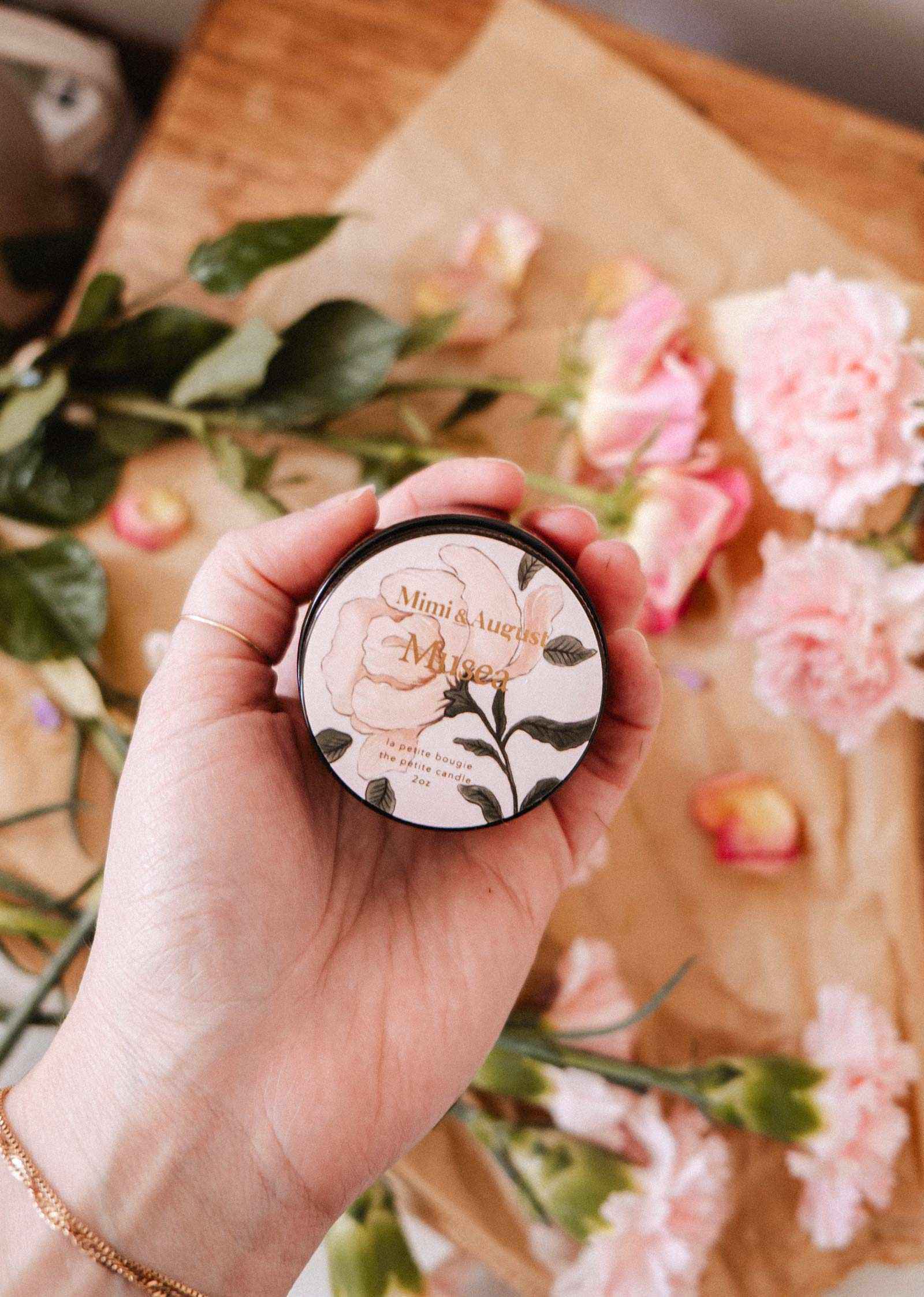 Une main tient une petite boîte ronde étiquetée « Mimi & August Musea », une bougie de soya réutilisable de 2 oz, avec des motifs floraux sur l'étiquette; des fleurs roses et des feuilles vertes décorent l'arrière-plan.