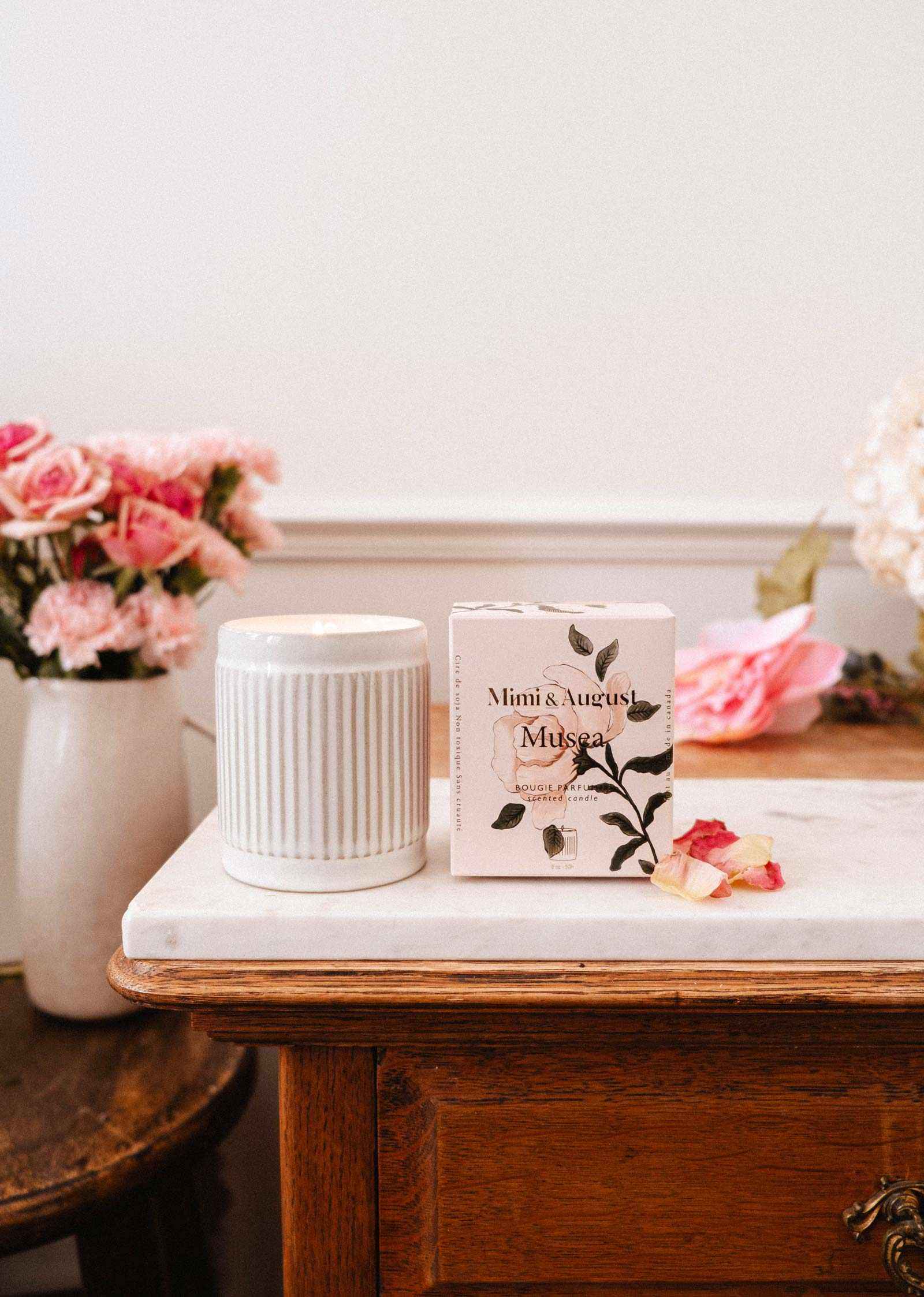 A Musea – Reusable Soy Candle by Mimi & August is displayed on a marble-topped wooden table with a vase of pink flowers in the background.