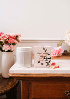 A Musea – Reusable Soy Candle by Mimi & August is displayed on a marble-topped wooden table with a vase of pink flowers in the background.