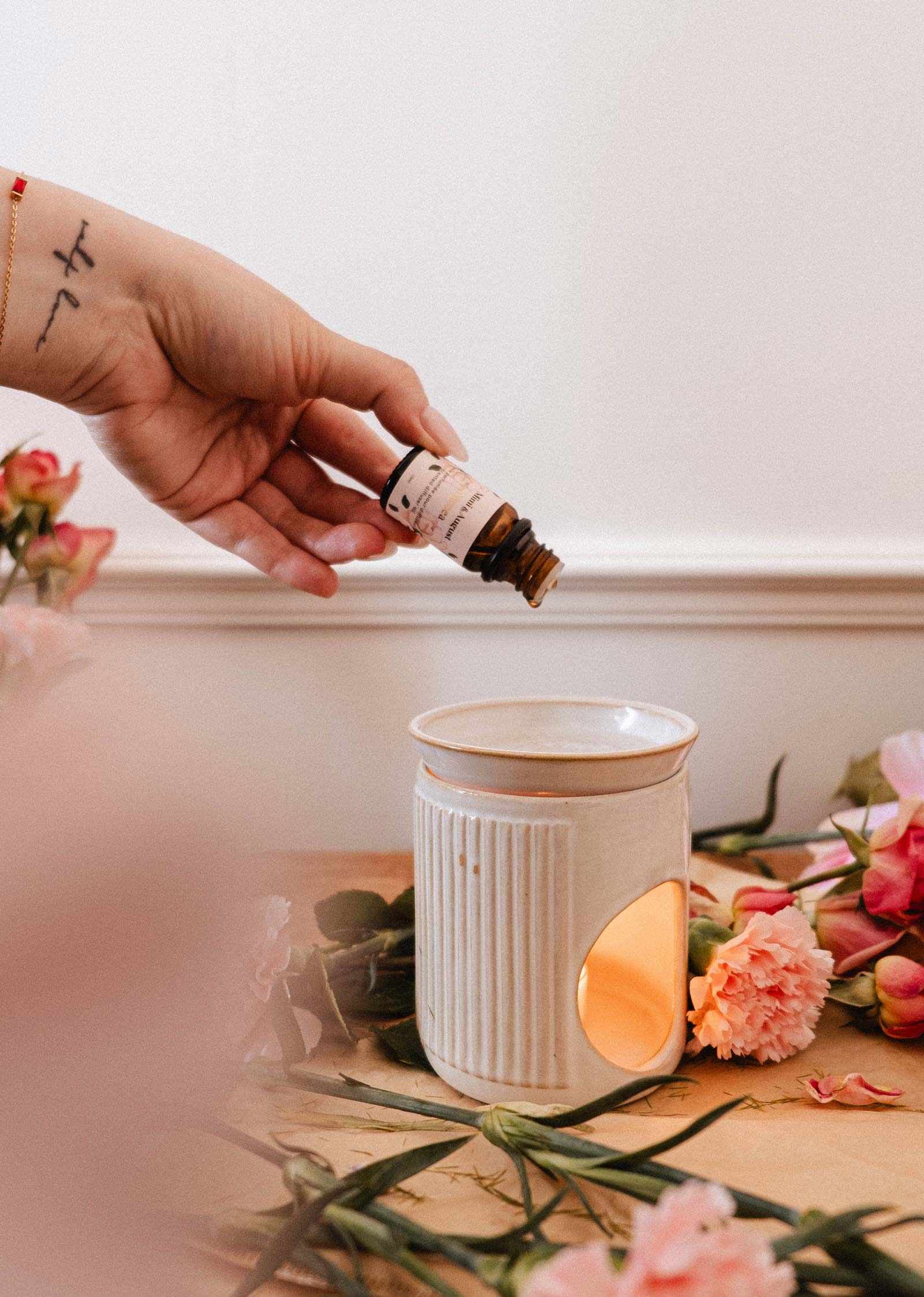 Une main verse l'huile parfumée Musea de Mimi & August, au parfum d'orange sanguine, dans un brûleur en céramique blanc entouré de fleurs roses — une charmante alternative au diffuseur ultrasonique.
