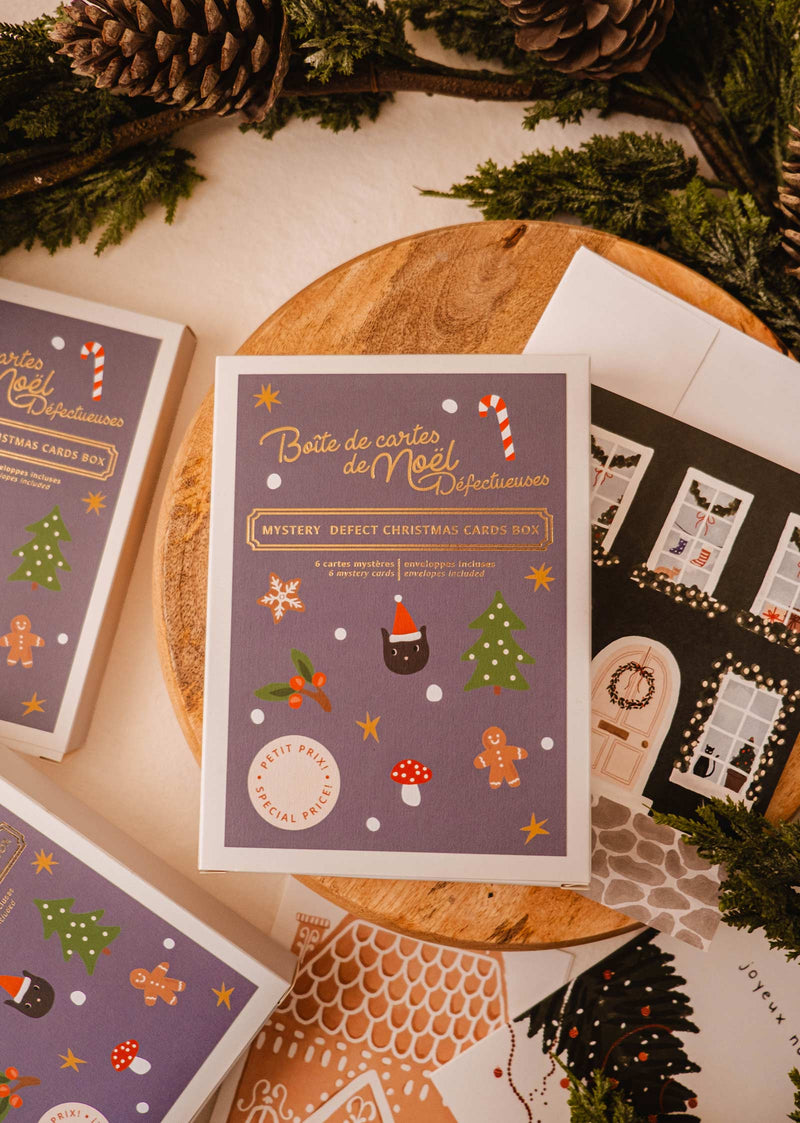 Embrace the holiday spirit with Mimi & August's Mystery Defect Christmas Card Box – Set of 6. This budget-friendly collection is elegantly adorned in purple, showcasing festive illustrations such as a cat in a Santa hat. It includes additional Christmas cards displayed on a wooden surface, surrounded by greenery.