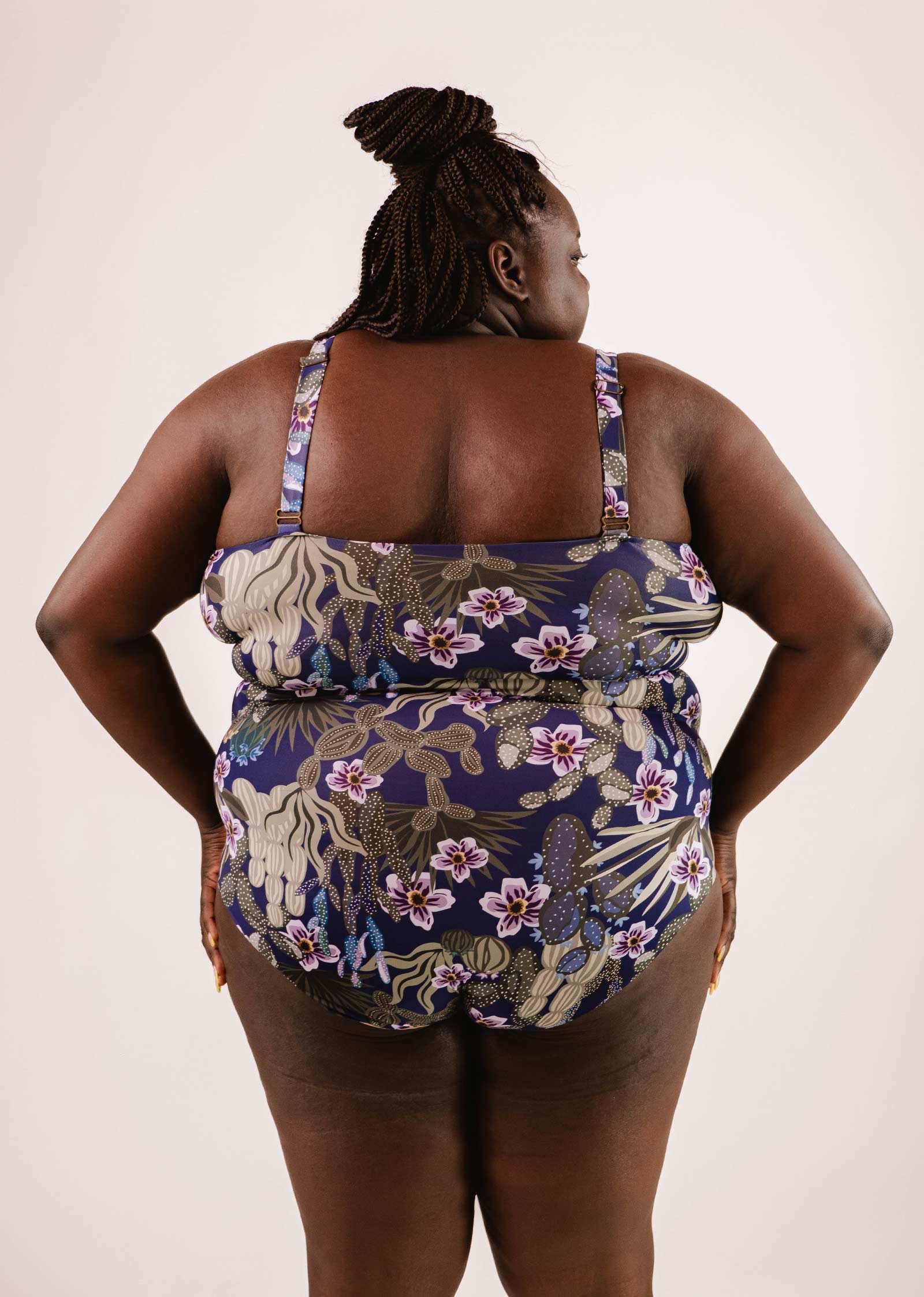 A person with dark braided hair, viewed from the back, wearing a Nohea Jardin de Nuit One-Piece Swimsuit by Mimi & August with a floral pattern in navy blue, beige, and purple colors. The swimsuit features adjustable straps and removable pads for a customizable fit.