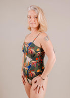 Mature woman with gray hair, wearing a colorful Mimi & August Nohea Tropica one-piece swimsuit with adjustable straps, standing confidently against a neutral backdrop.