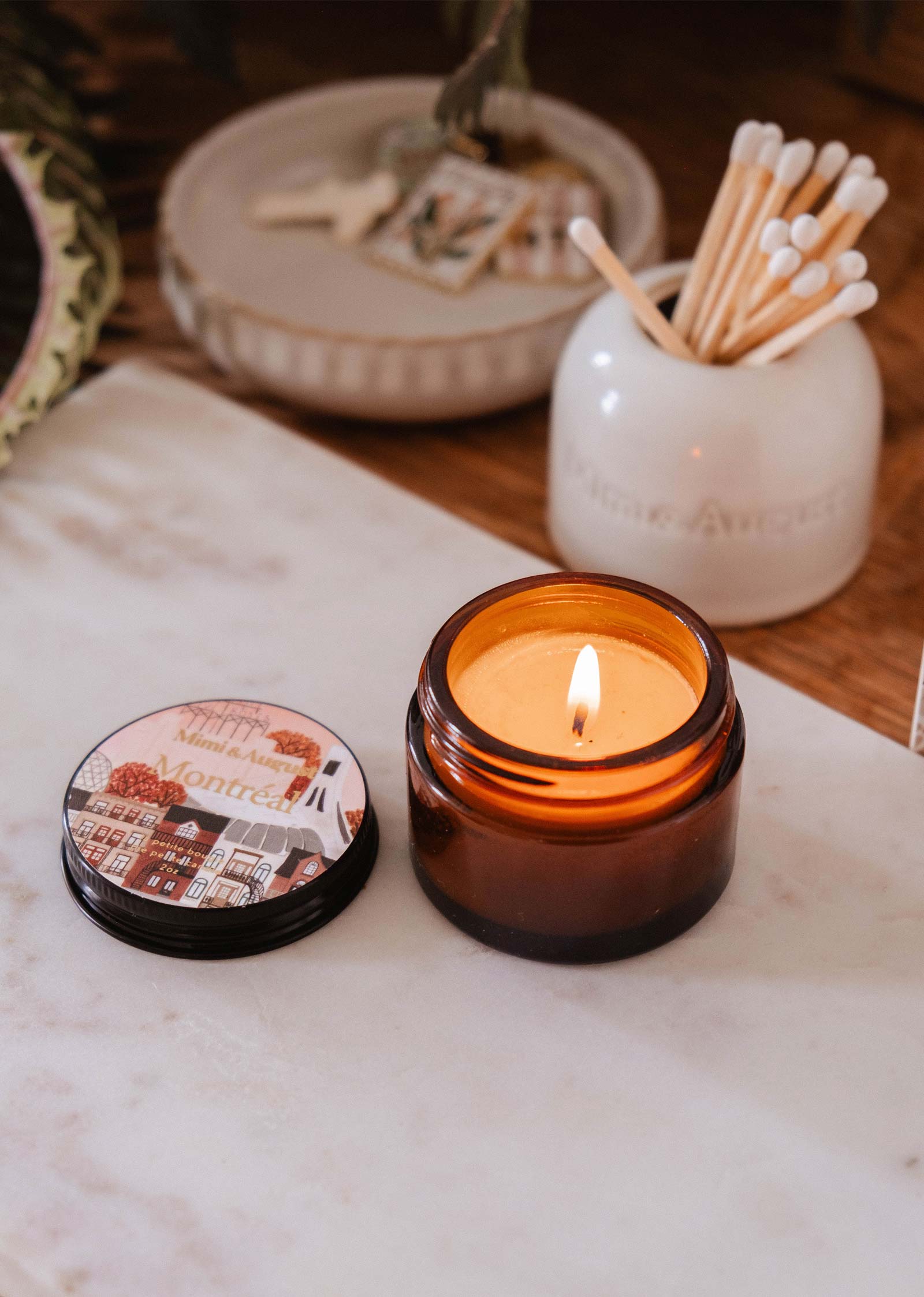 The Mimi & August Montreal - 2 oz candle, in a small amber glass jar with a dark rose scent, sits lit beside its illustrated lid and a swab-filled ceramic holder. This reusable candle brings elegant fragrance to any room.