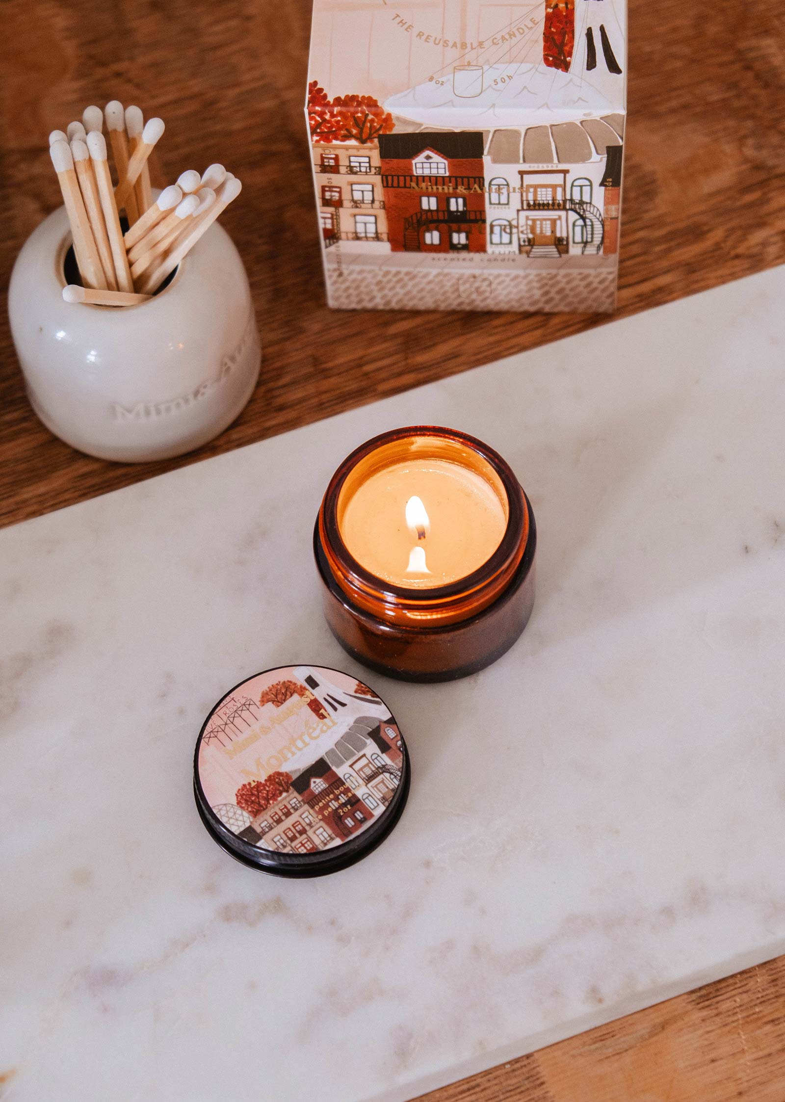 A Mimi & August Montreal - 2 oz candle with a dark rose scent sits in a brown glass jar on a marble tray next to its illustrated lid, with a jar of matches and a decorative box visible in the background.