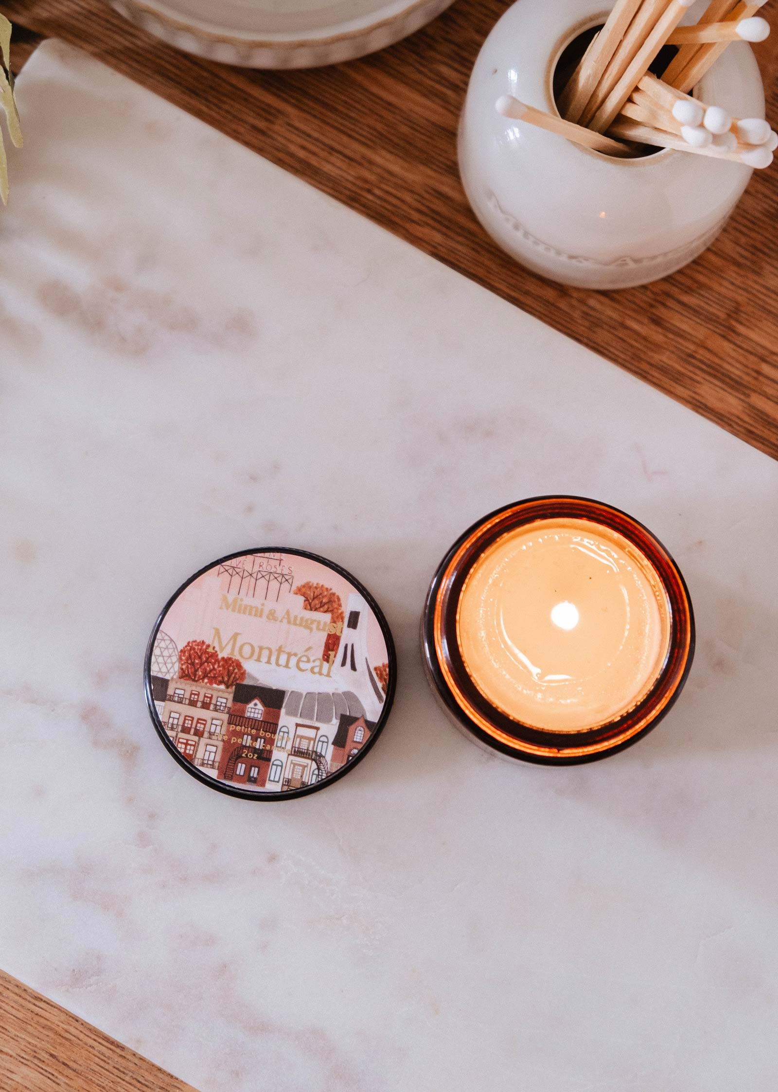A 2 oz Montreal candle by Mimi & August with a dark rose scent in an amber jar sits on marble near a city-illustrated lid, flanked by cotton swab and match jars.