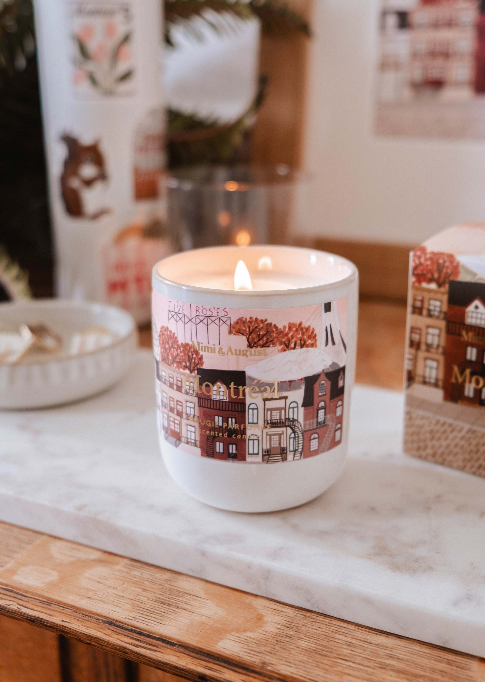 The Montreal - Reusable Candle by Mimi & August, a lit scented candle in a decorative white jar with illustrated buildings, sits on a marble tray atop a wooden surface, surrounded by small objects.