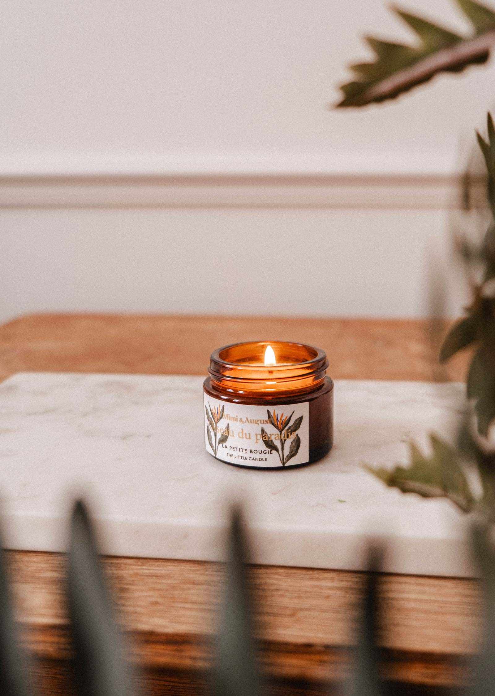 A lit Mimi & August Oiseau du Paradis – 2 oz Soy Candle in an amber glass jar rests on a white marble slab atop wood, with green leaves in the foreground.