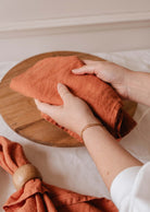 Une personne plie un linge en lin Mimi & August, ressemblant à une serviette, sur une planche ronde en bois, avec un autre torchon et un rond de serviette reposant sur une nappe blanche à proximité.