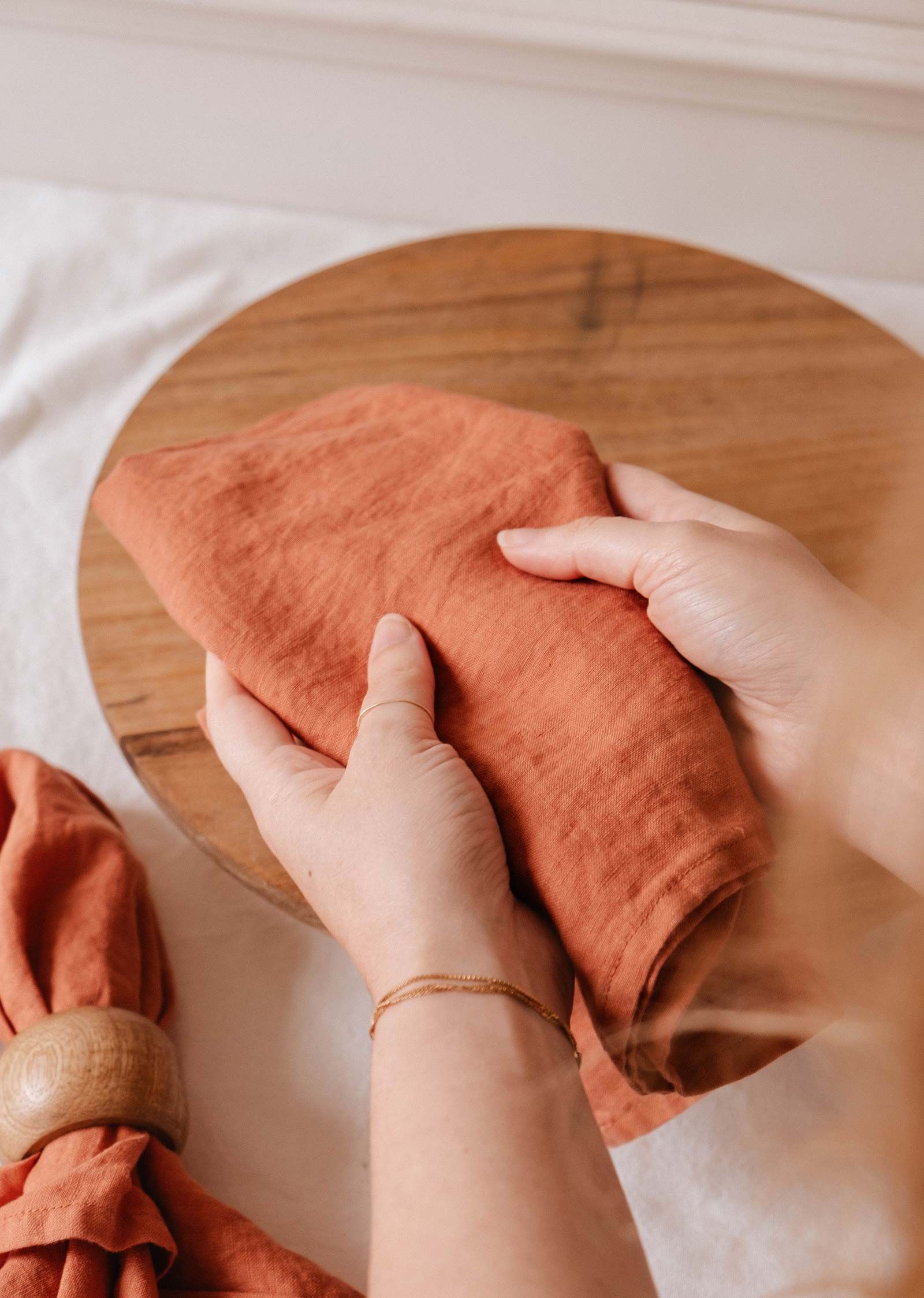 Une personne plie le linge en lin de couleur rouille de Mimi & August sur une planche ronde en bois, avec une serviette et un anneau à proximité. Idéal pour sécher la vaisselle ou ajouter une touche d'élégance à votre table.