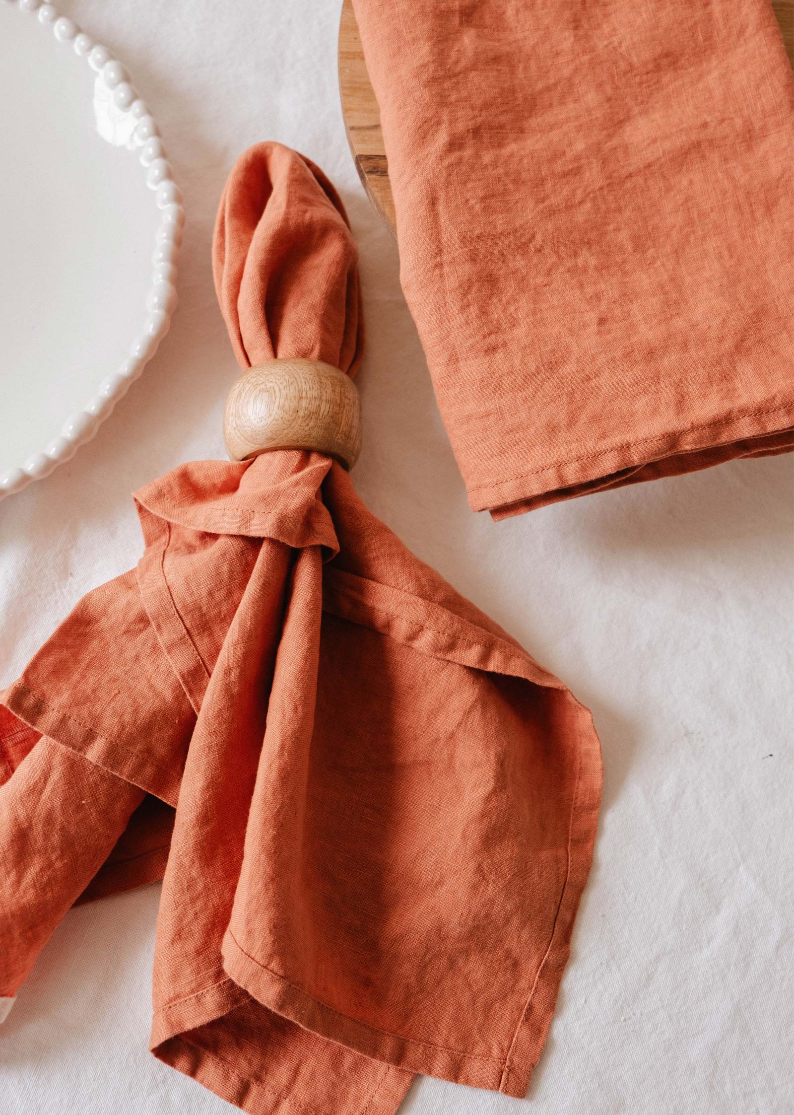 Un linge en lin doux de couleur rouille de Mimi & August, associé à une serviette en lin assortie avec un anneau en bois, se trouve à côté d'une assiette blanche à bord perlé sur une nappe blanche.