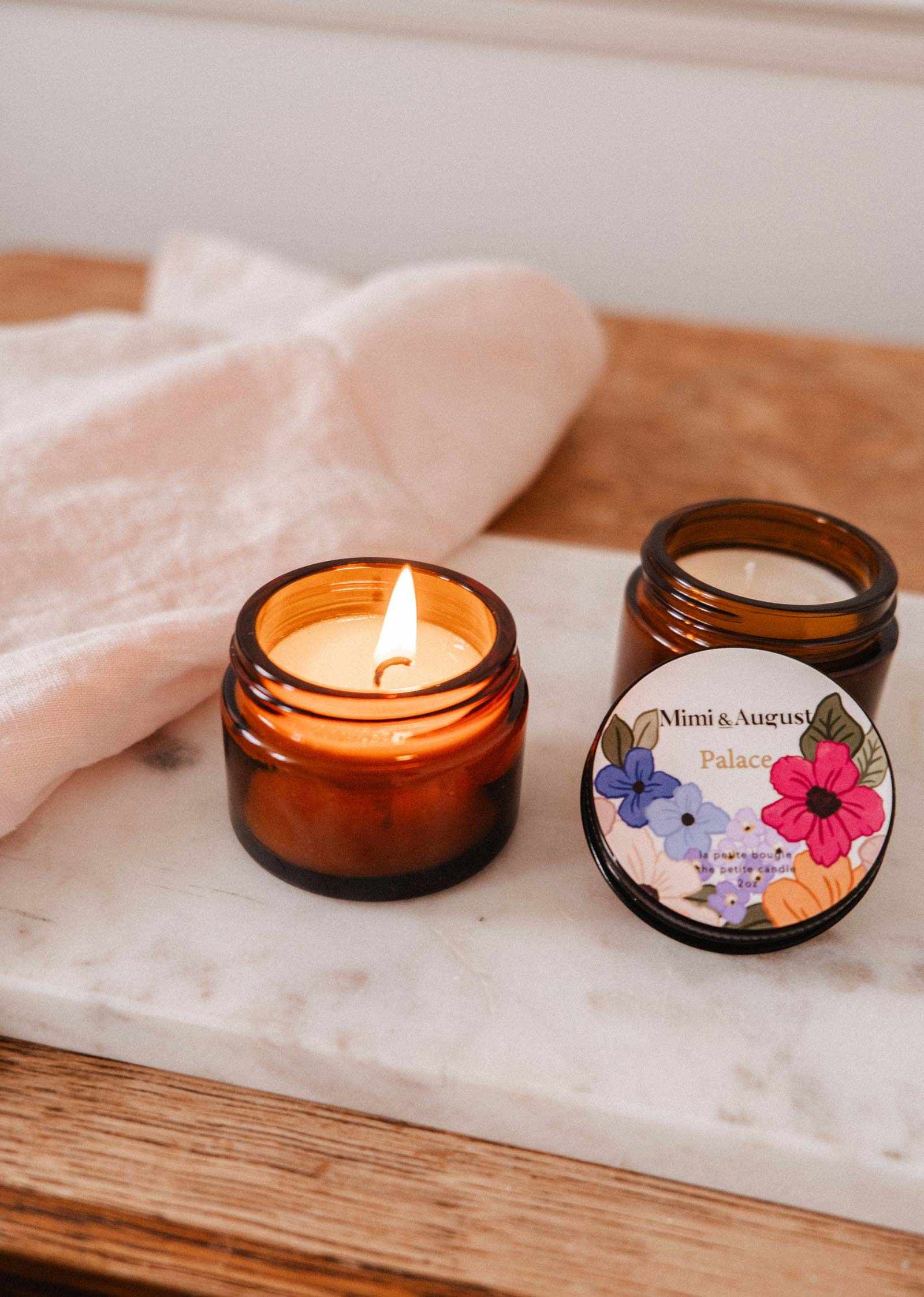 Two Mimi & August Palace 2 oz soy candles in brown glass jars sit on a marble tray; one is lit, the other has its floral lid off, revealing its branded label.