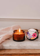 La bougie de soya Palace 2 oz de Mimi & August, coulée à la main à Montréal, repose dans un pot en verre brun sur du marbre à côté de son couvercle floral et d'un tissu rose pâle plié.