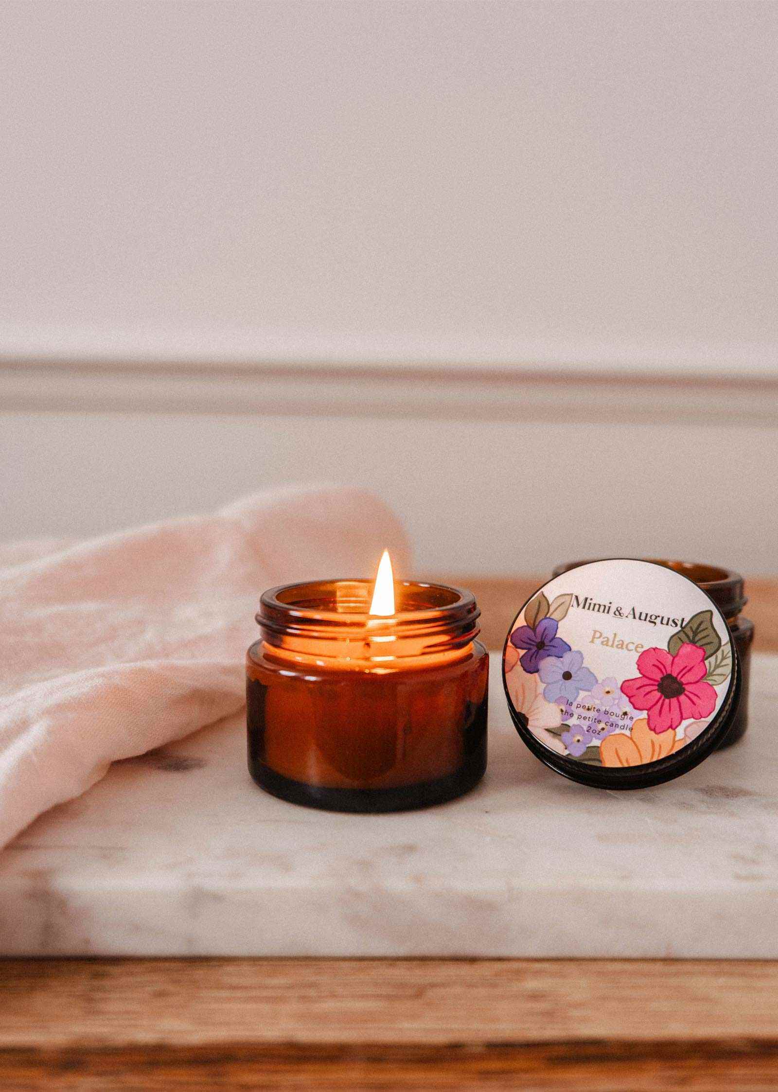 The Palace 2 oz soy candle by Mimi & August, hand-poured in Montreal, sits in a brown glass jar on marble next to its floral lid and a folded light pink cloth.