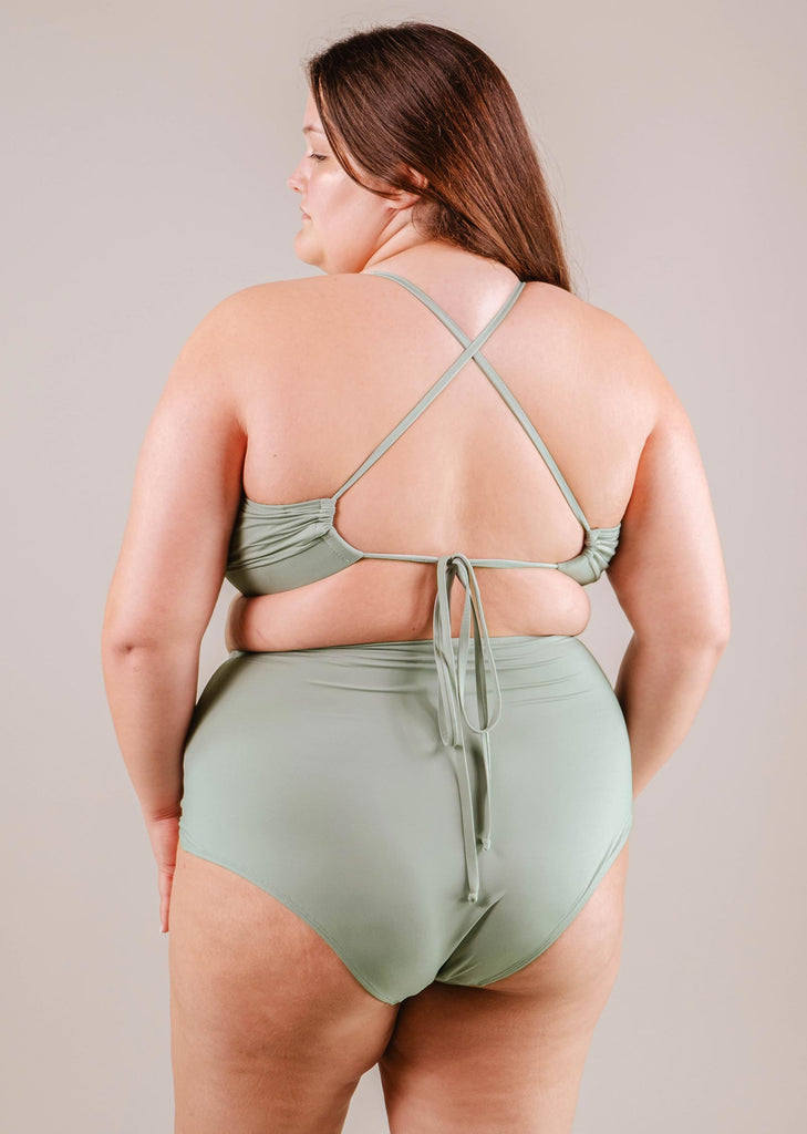 Une femme vue de dos, portant un bas de bikini taille haute Paloma Agave de Mimi & August, debout sur un fond neutre.