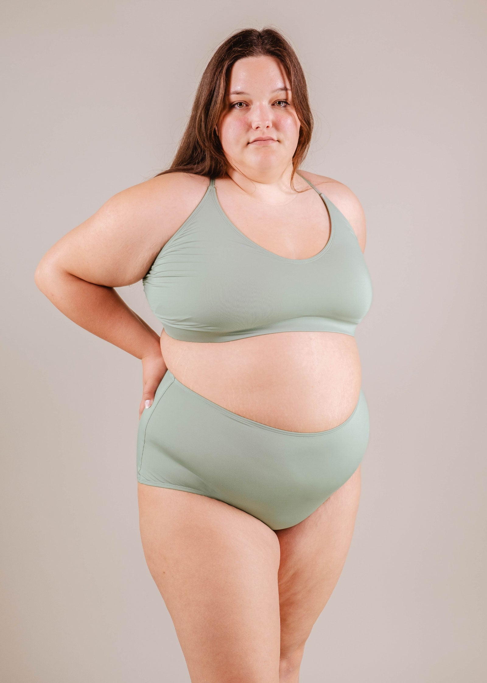 A woman in a Paloma Agave High Waist Bikini Bottom and Mimi & August brand stands confidently, one hand on her hip, against a neutral background.