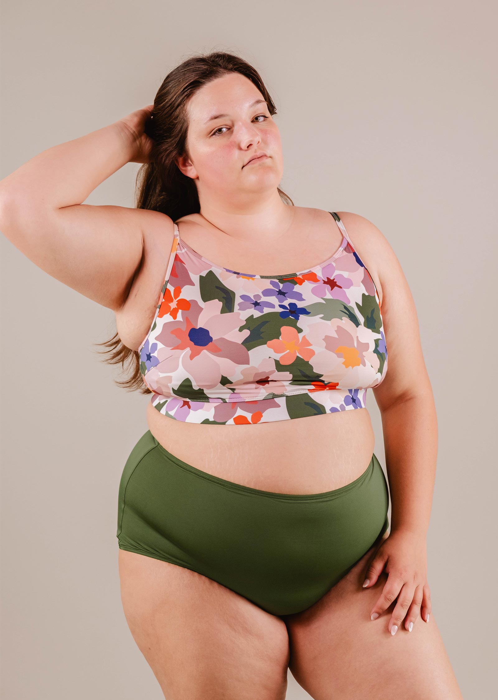 Un mannequin grande taille vêtu d'un débardeur à fleurs et d'un bas de bikini taille haute Paloma Amazonia de Mimi & August, posant avec une main sur la hanche et l'autre dans les cheveux, sur un fond neutre.