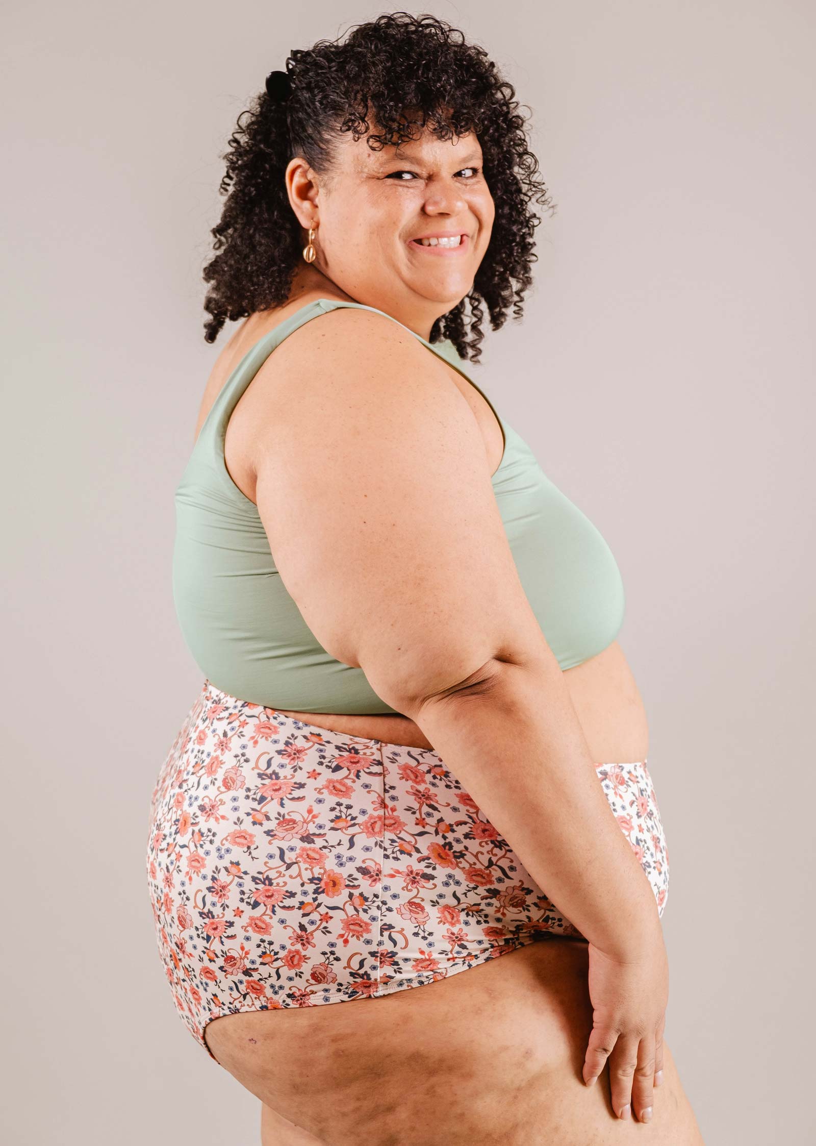 A woman with curly hair smiling at the camera, wearing a mint green tank top and Mimi & August's Paloma Amour High Waist Bikini Bottom, standing in a neutral-colored studio.