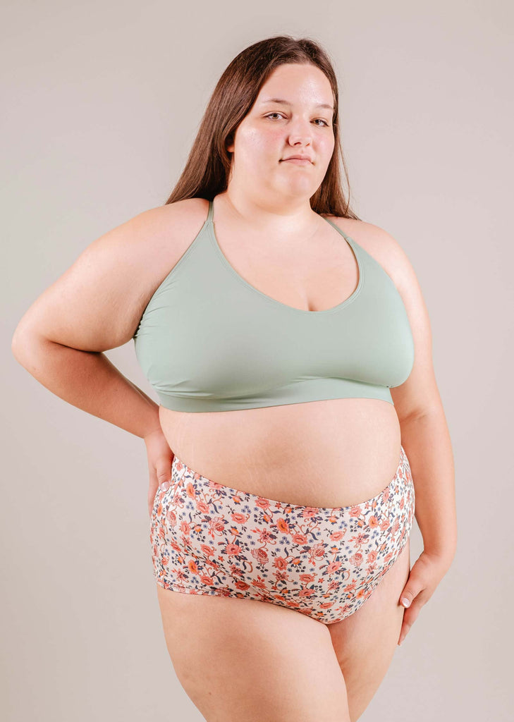 A confident plus-size woman in a green sports bra and Paloma Amour High Waist Bikini Bottom shorts standing against a neutral background by Mimi & August.