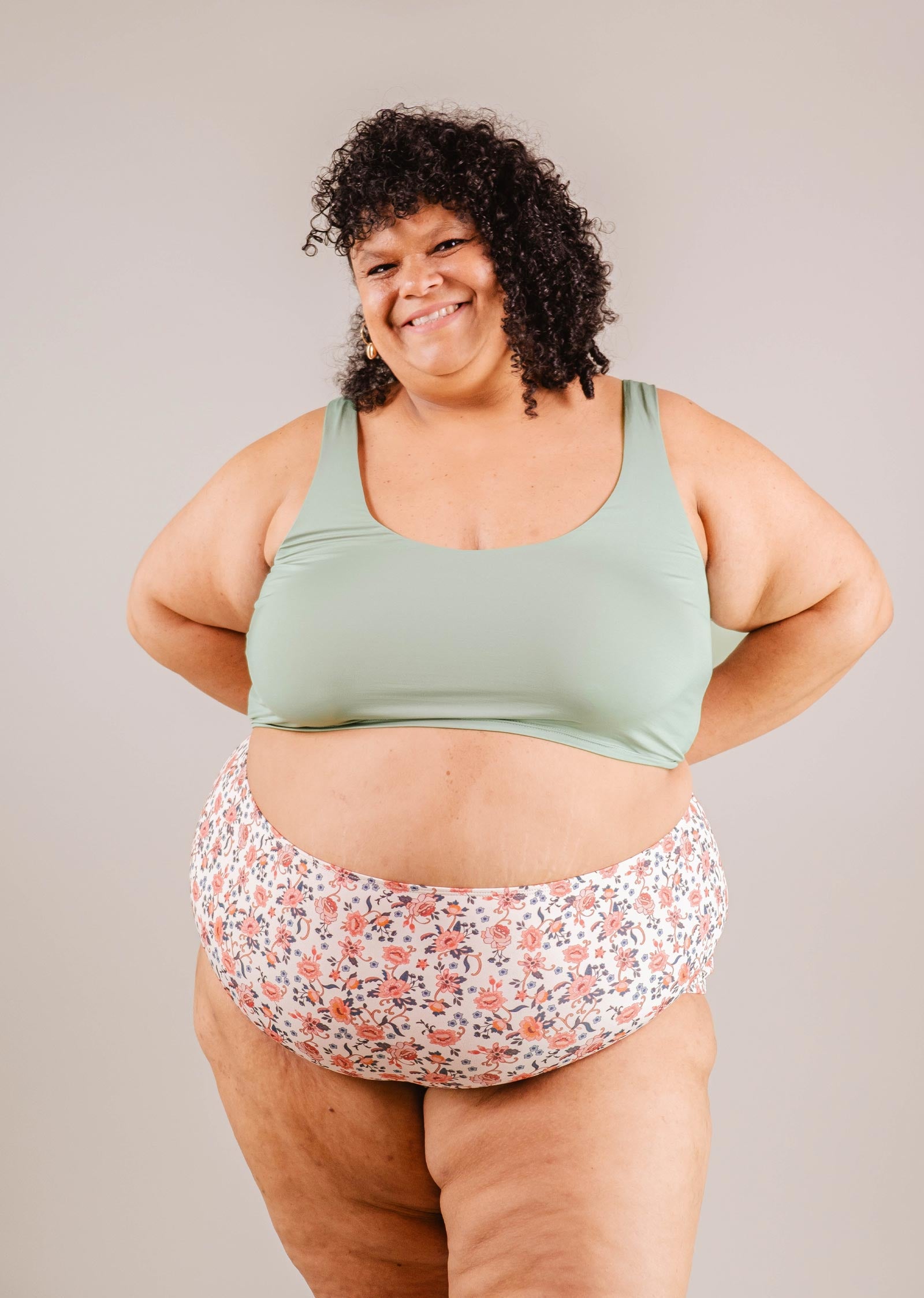 A cheerful woman in a green sports bra and Paloma Amour High Waist Bikini Bottom by Mimi & August, standing confidently with hands on hips against a neutral background.