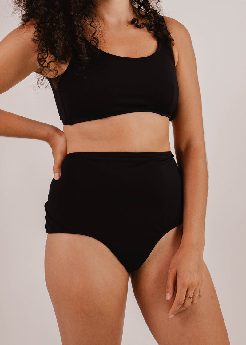Person wearing a black two-piece swimsuit, featuring the Paloma Black High Waist Bikini Bottom by Mimi & August, with one hand resting on their hip against a plain background.