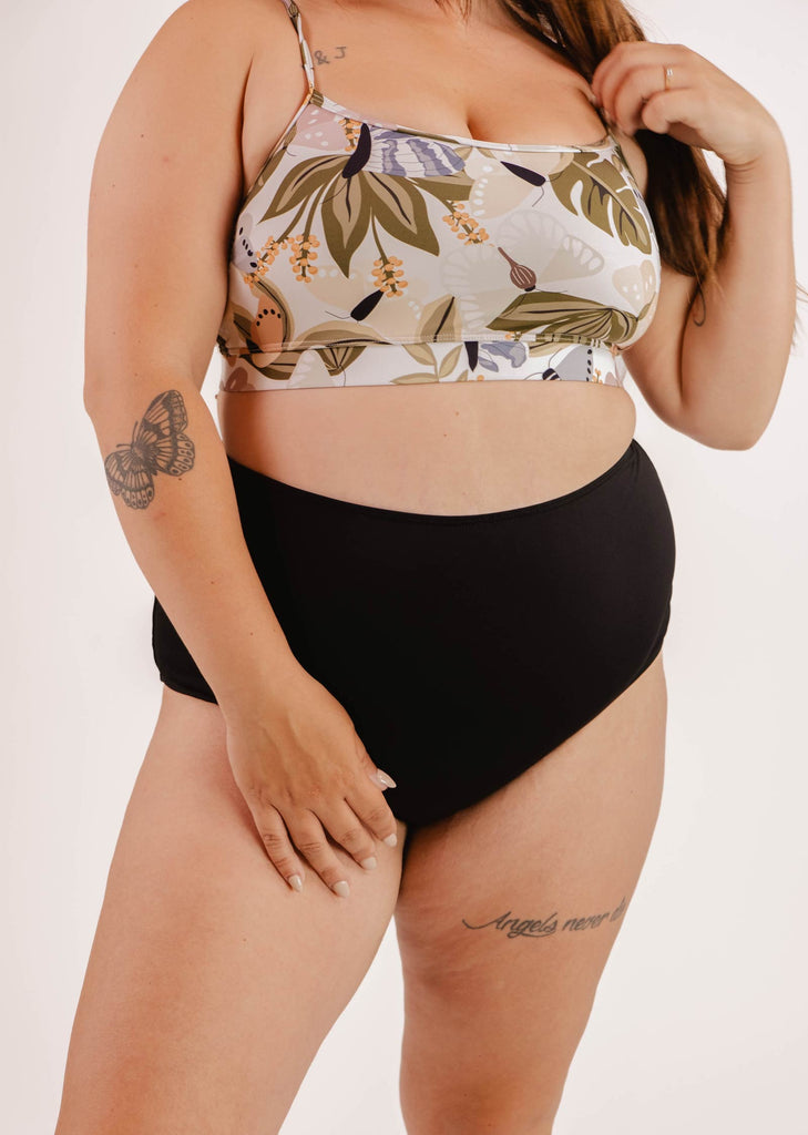 A person wearing a floral crop top and the Mimi & August Paloma Black High Waist Bikini Bottom, showing their tattoos on the arm and thigh.