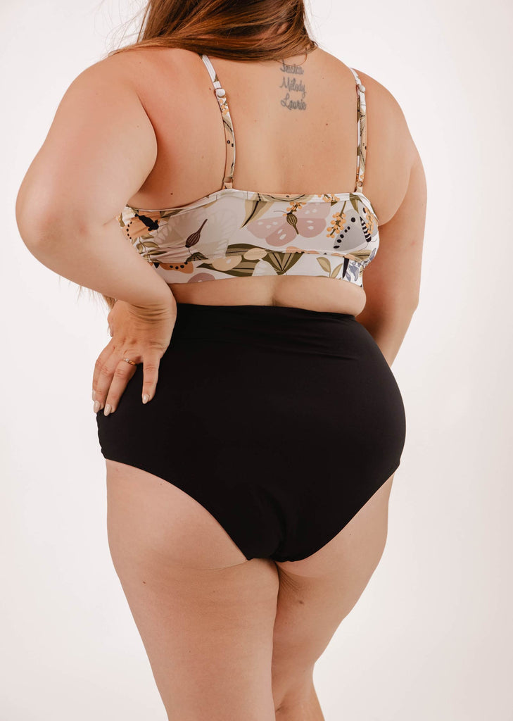 Une personne se tient debout, dos à la caméra, vêtue d'un haut de bikini à fleurs et d'un bas de bikini taille haute noir Mimi & August Paloma, un véritable élément de base des vêtements de plage, avec un tatouage sur le haut du dos.