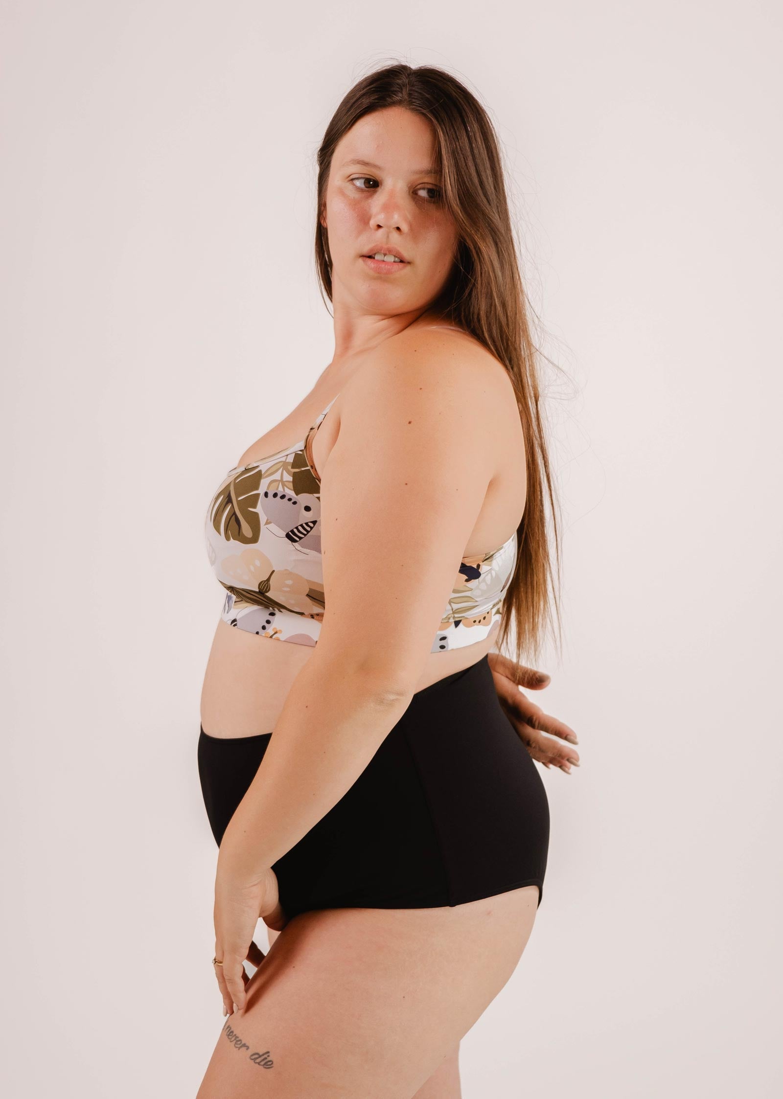 A woman with long hair stands sideways, wearing a patterned sports bra and Mimi & August Paloma Black High Waist Bikini Bottom against a plain background.