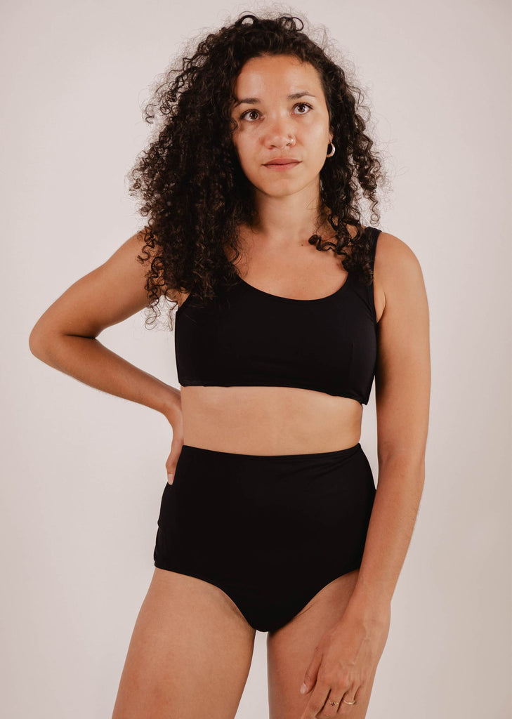 A woman with curly hair is wearing a black two-piece swimsuit. She is standing with one hand on her hip, looking slightly off camera against a plain background. The high-waisted swimwear style of her Paloma Black High Waist Bikini Bottom from Mimi & August exudes timeless elegance and confidence.