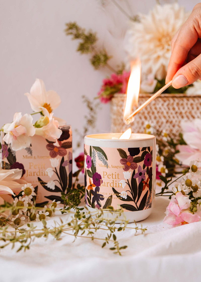 Une main allume une bougie réutilisable Petit Jardin avec une allumette, entourée de fleurs fraîches et de verdure. Deux bougies similaires de Mimi & August sont placées à l'arrière-plan, emplissant l'air d'un parfum serein.