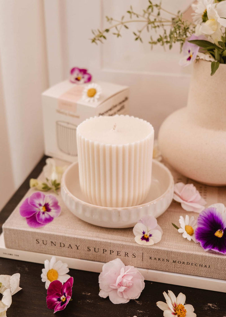 A white ribbed candle refill called "Candle Refill - Petit Jardin" by Mimi & August with an enchanting scent sits in a bowl on a book titled "Sunday Suppers". Flowers are scattered around the candle, and a vase of flowers is seen in the background.