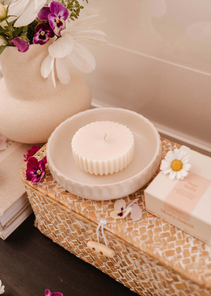 A round white candle refill - Petit Jardin by Mimi & August with an enchanting scent in a ceramic dish sits on a woven basket with a button clasp. An eco-friendly white vase with purple and white flowers is nearby, along with a box on top of books.