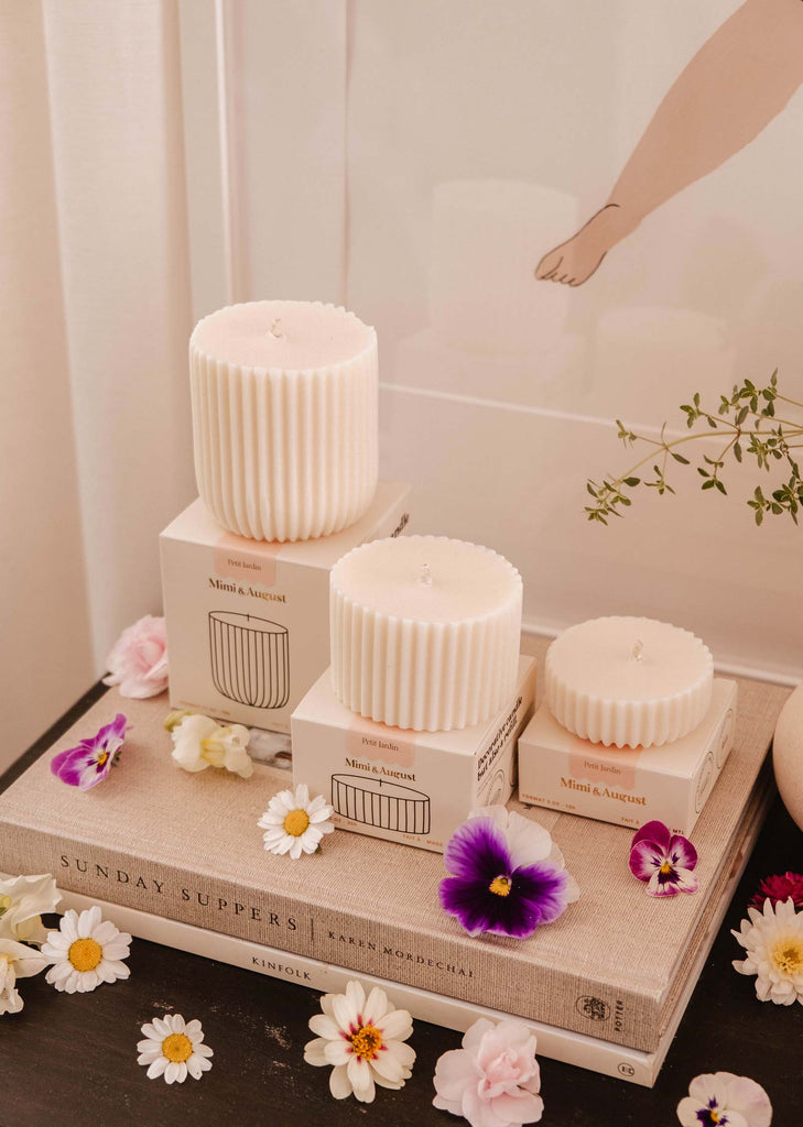 Three Candle Refill - Petit Jardin by Mimi & August in varying sizes sit on branded boxes atop a book, surrounded by flowers. An eco-friendly framed art piece and a small plant are in the background, enhancing the enchanting scent of the scene.