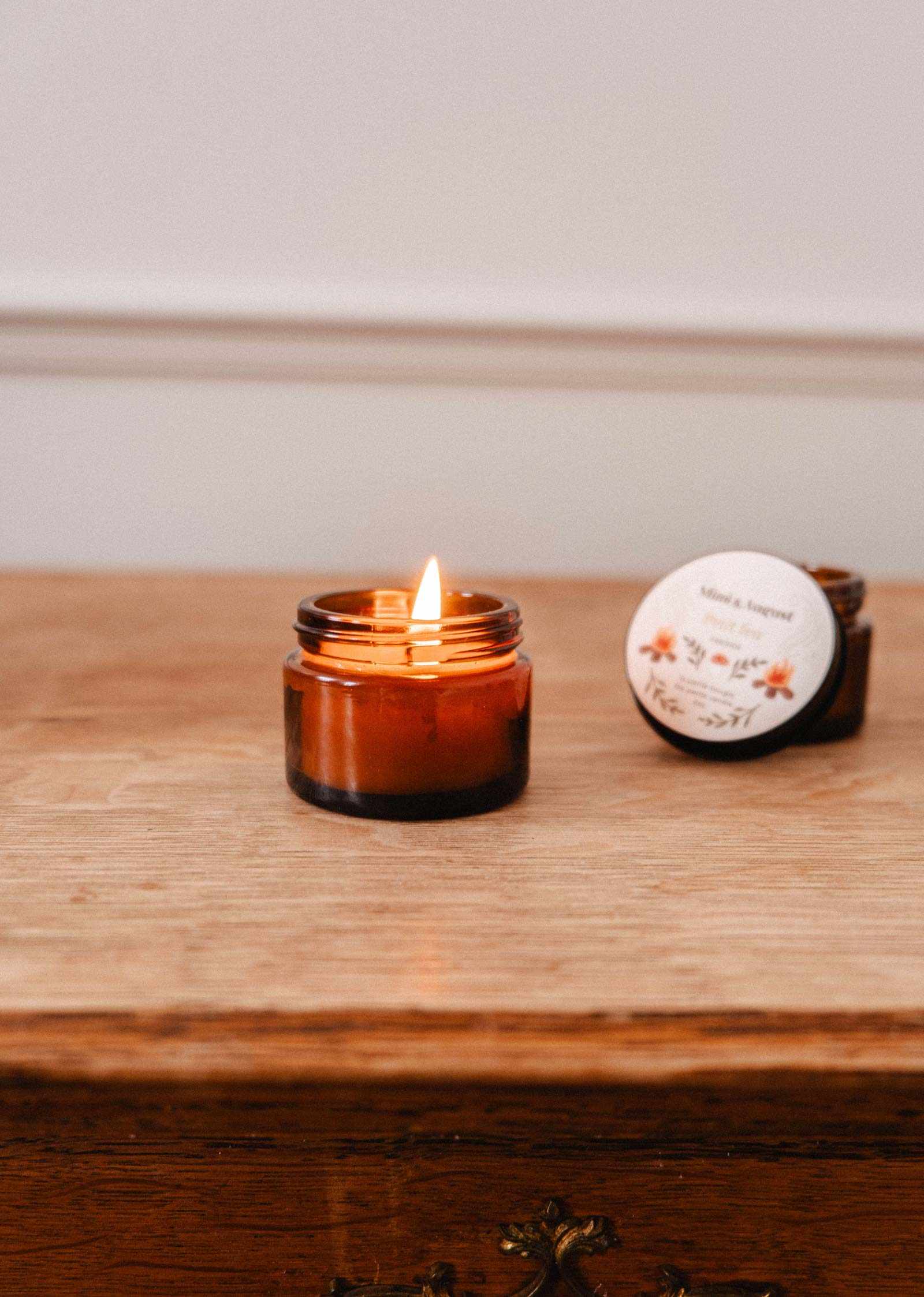 A lit Petit Feu – 2 oz Soy Candle by Mimi & August sits on a wooden surface, its labeled lid with floral graphics nearby. This cozy brown glass soy candle is hand-poured in Montreal.