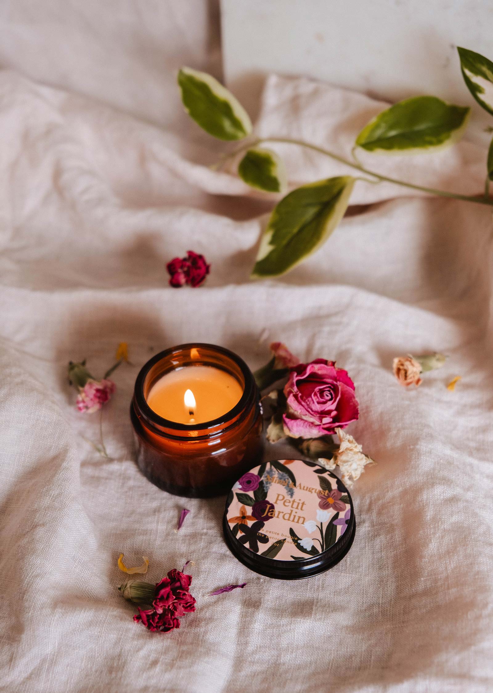The Petit Jardin - 2 oz candle by Mimi & August rests in a brown glass jar on fabric beside its floral lid, dried flowers, and green leaves, filling the space with a fresh water lily aroma for a tranquil vibe.