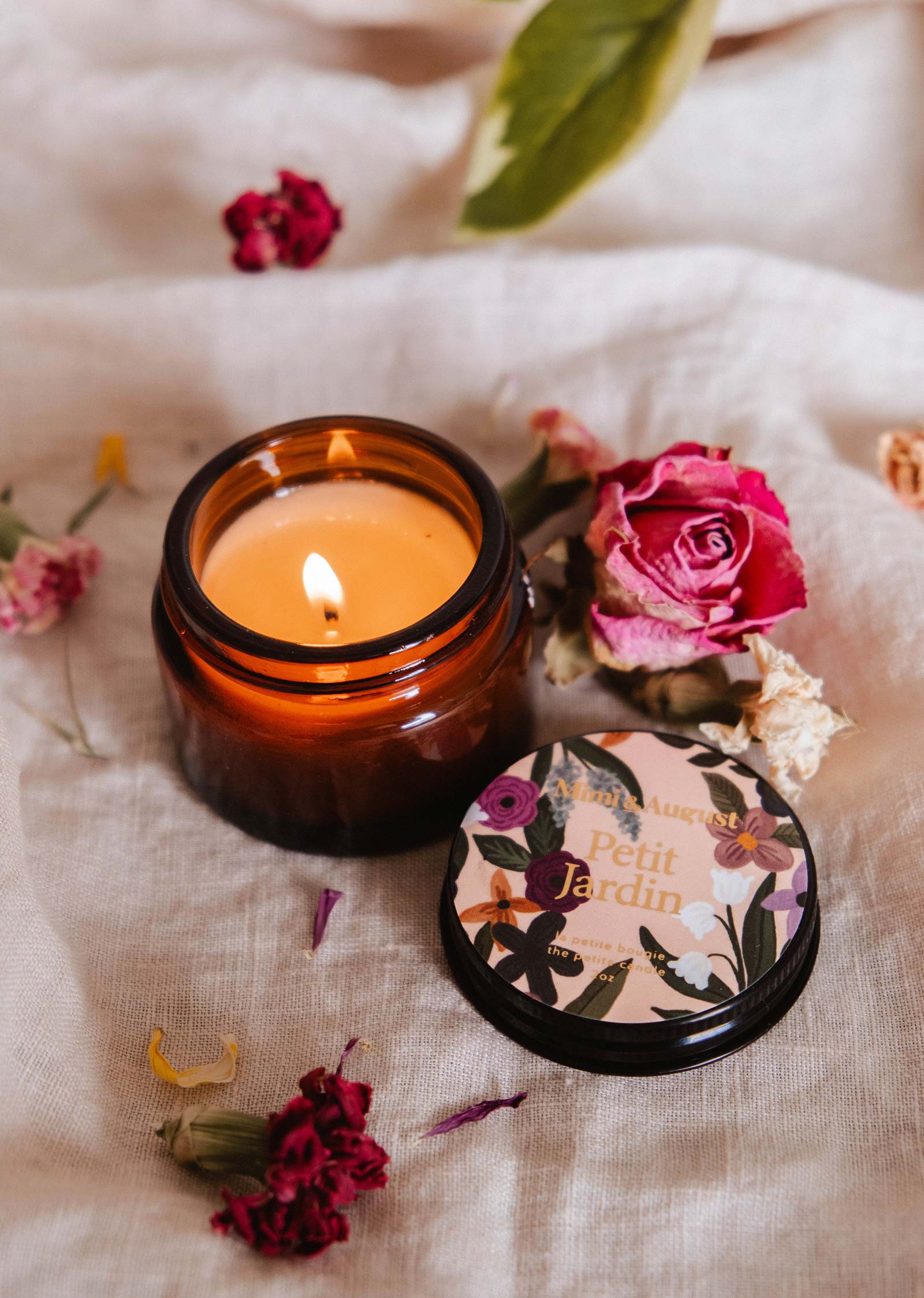A lit Mimi & August Petit Jardin - 2 oz candle glows in a brown glass jar on light fabric, with dried flowers around it. Its labeled lid nearby hints at water lily freshness, creating a tranquil oasis.