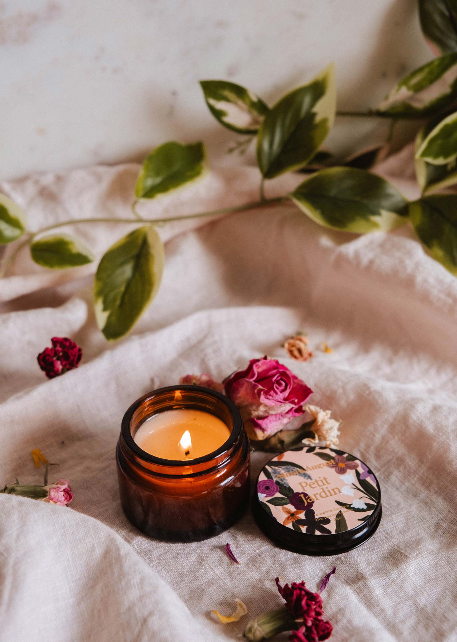 A lit Petit Jardin - 2 oz candle by Mimi & August, in a brown glass jar with a floral lid, sits on soft beige fabric among dried flowers and leafy branches, evoking the fresh scent of water lilies.