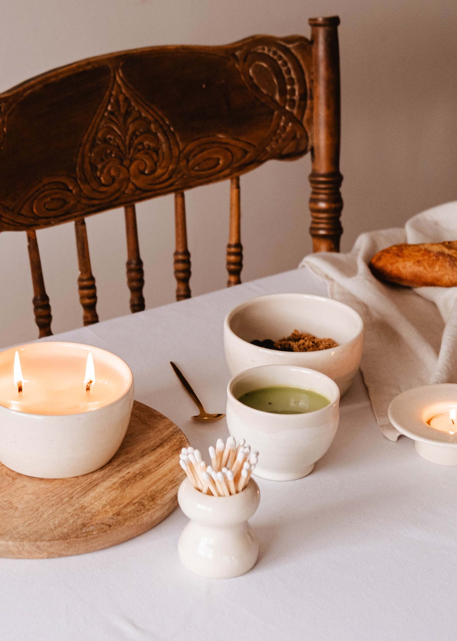 La table est décorée d'une bougie allumée, de petits bols de matcha et de granola, d'un pain posé sur une nappe blanche et de la Petite Flamme - Pot d'allumettes en céramique de Mimi & August, élégamment placée près de la chaise en bois. Des cotons-tiges complètent cette présentation artisanale.