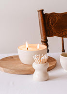 A lit candle in a ceramic holder sits on a wooden tray alongside the Mimi & August Petite Flamme - Ceramic Match Pot. Nearby, a small container of cotton swabs and a wooden chair complete the scene, highlighting artisanal home decor at its finest.