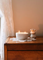 A lit candle in a ceramic bowl adorns the wooden dresser alongside an artisanal home decor piece, the Petite Flamme - Ceramic Match Pot by Mimi & August, while lace drapery elegantly frames the scene.